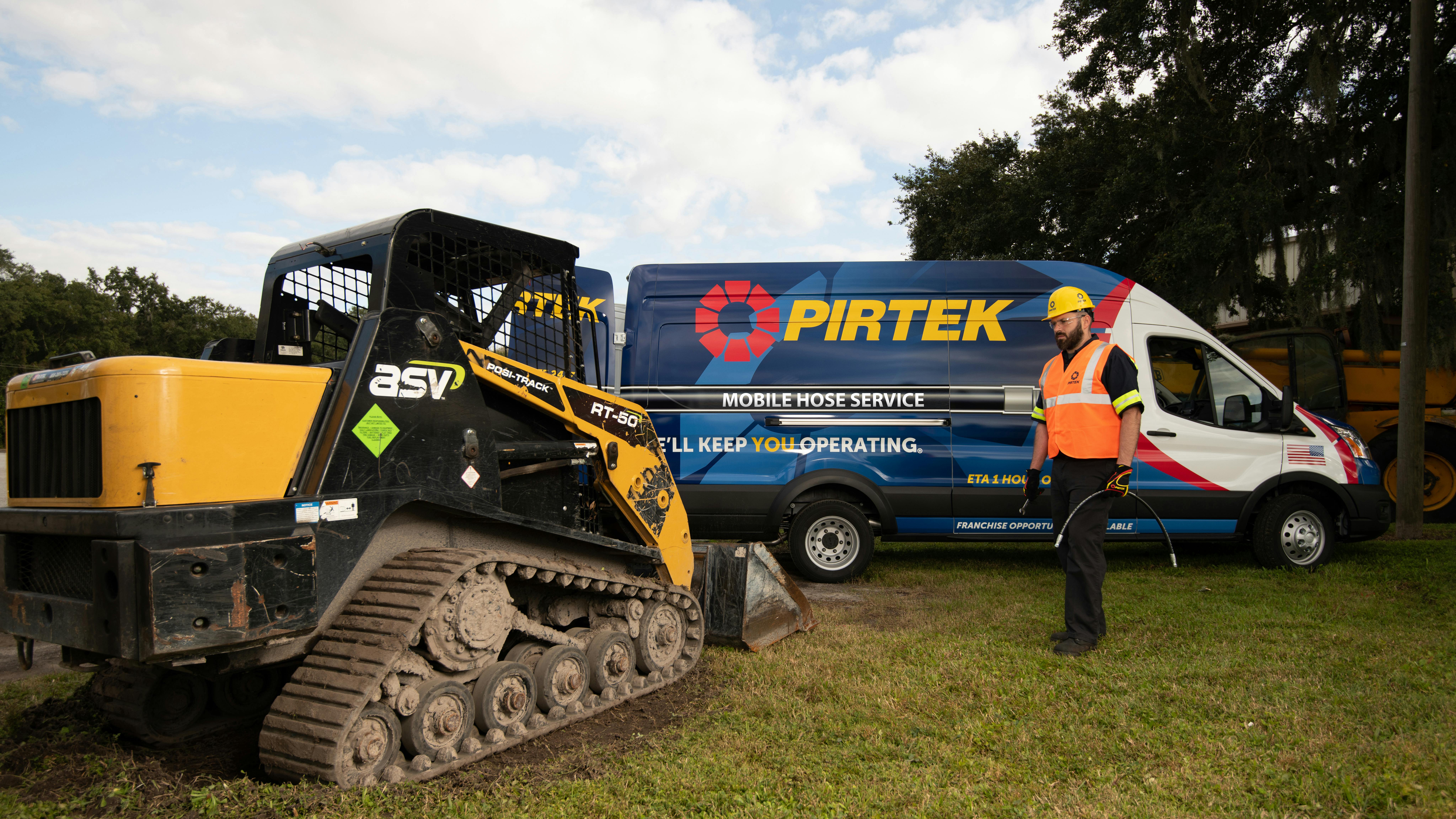 A PIRTEK technician holding a hydraulic hose walking toward a compact track loader